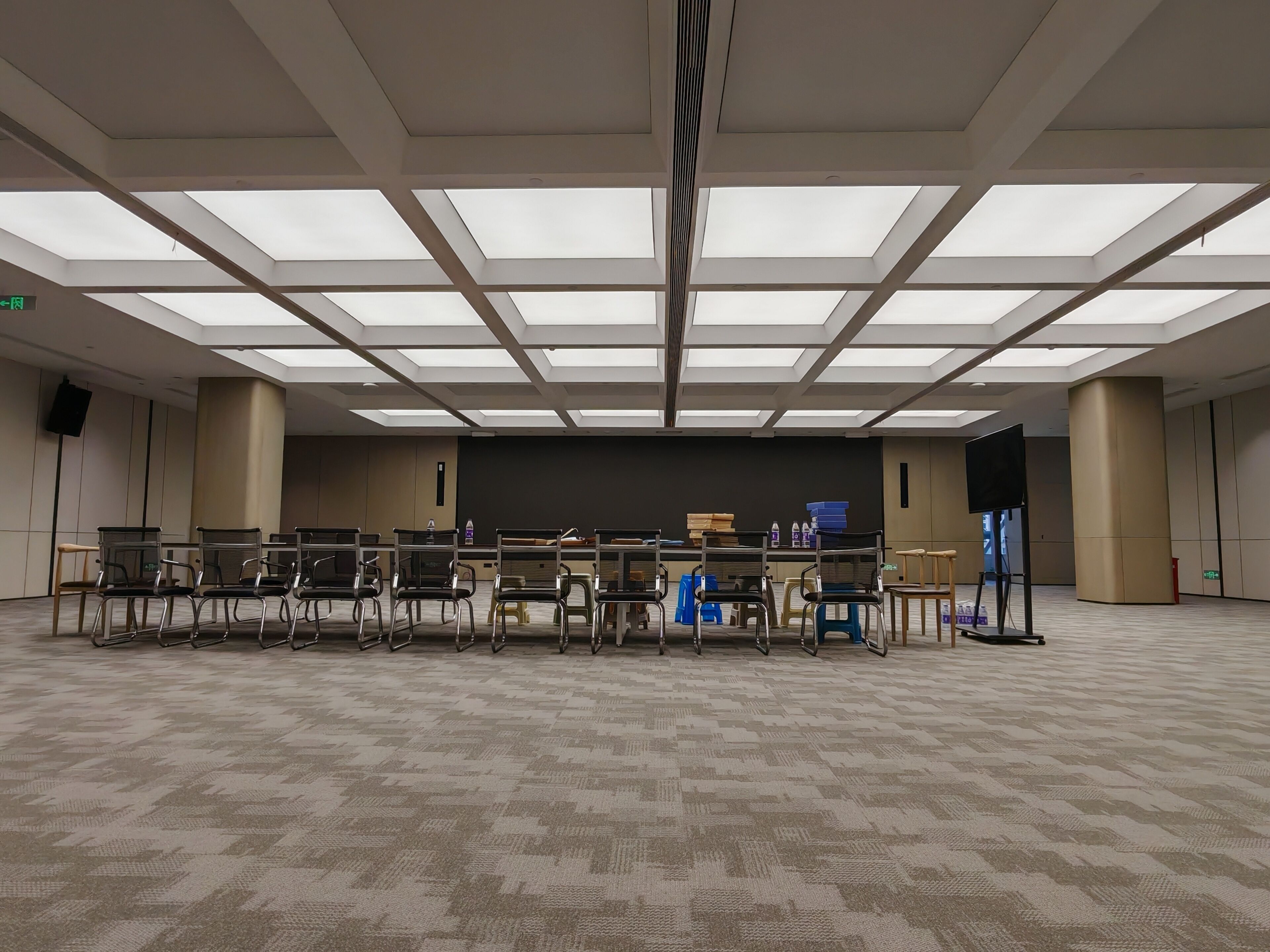 A conference room with a high ceiling, multiple rows of chairs, a stage, and a large screen.