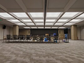 Un salón de conferencias con techo alto, varias filas de sillas, un escenario y una pantalla grande.