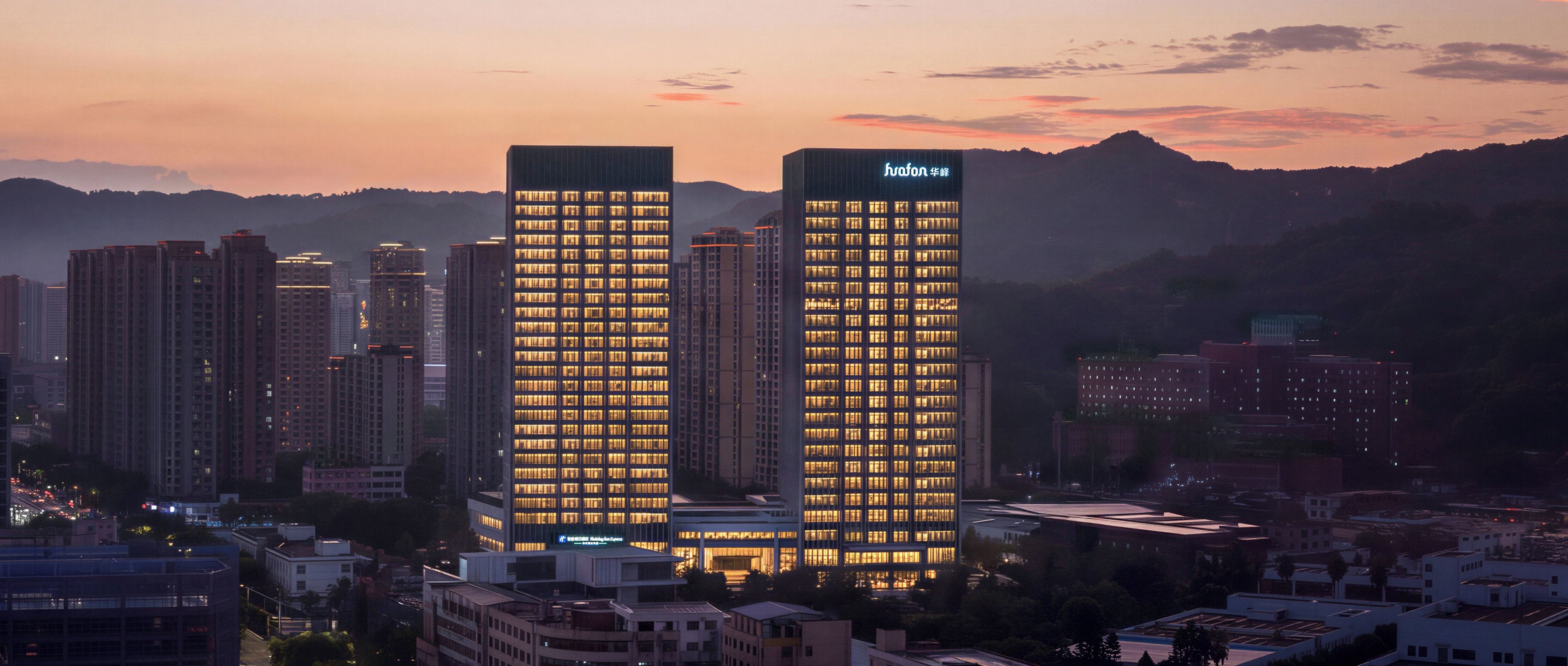 A cityscape at dusk with two prominent high-rise buildings, one with illuminated windows and a sign, and the other with a neon sign.