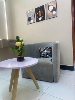 A room with a grey sofa, a white round table with a vase of yellow flowers, and three wall-mounted light fixtures. - JS AIRBNB APARTMENTS
Luxury rooms that feel like home’ (Kampala)