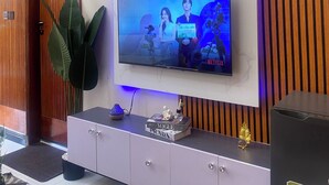 Uma sala de estar moderna com uma TV de tela plana fixada numa parede branca, um painel de madeira e um armário cinza com uma pequena planta, além de um alto-falante preto.
