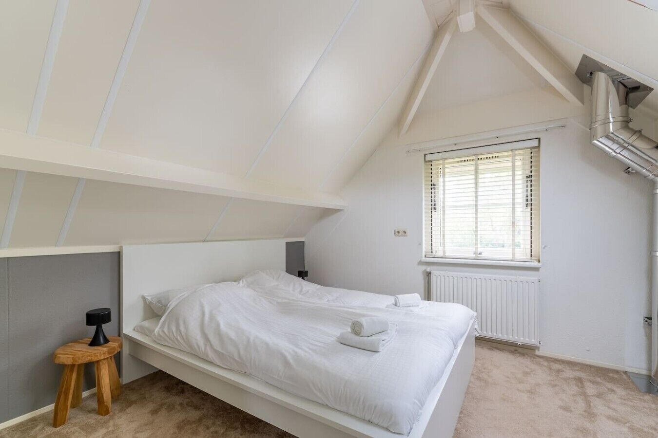 A bedroom with a sloped ceiling, a bed, a window with blinds, a radiator, and a small wooden stool.