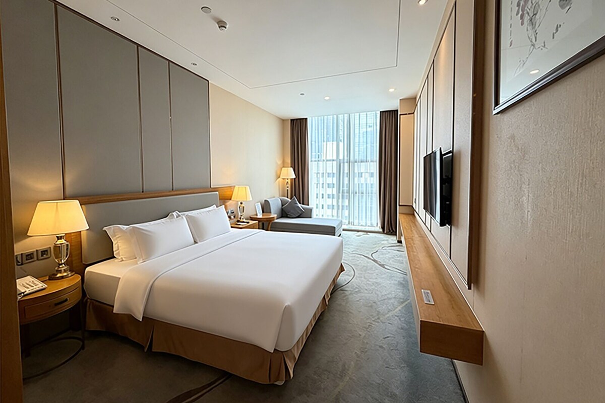 Een hotelkamer met een groot bed, nachtkastjes, een bureau, een stoel, een televisie en een raam met uitzicht op de stad.