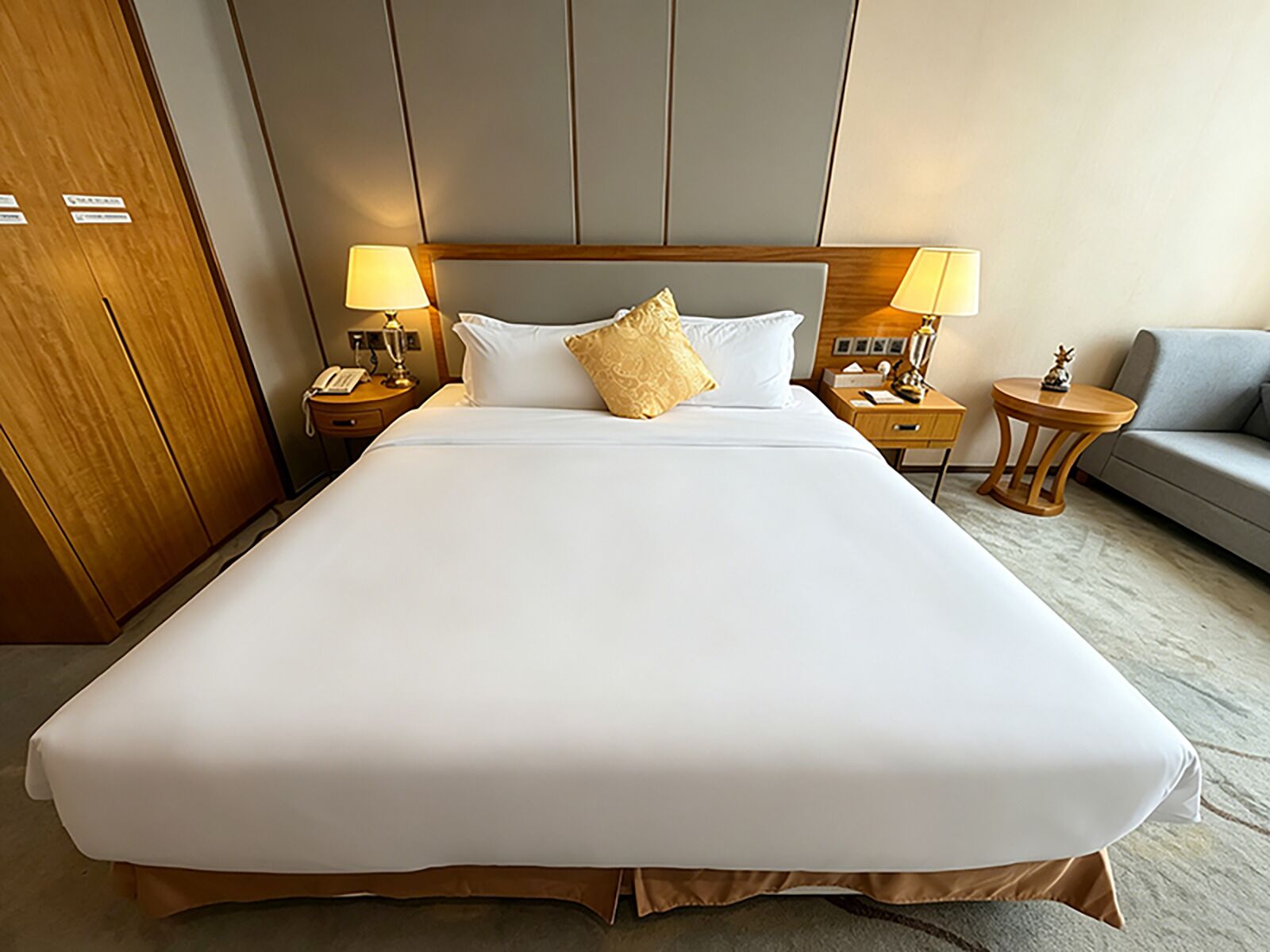 Una cama grande con sábanas blancas, dos mesitas de noche con lámparas, un cabecero de madera, un sofá gris y un armario de madera.