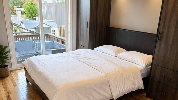 Une chambre avec un grand lit, un plancher en bois, un luminaire au plafond et une vue sur l’extérieur, accessible par une porte coulissante.