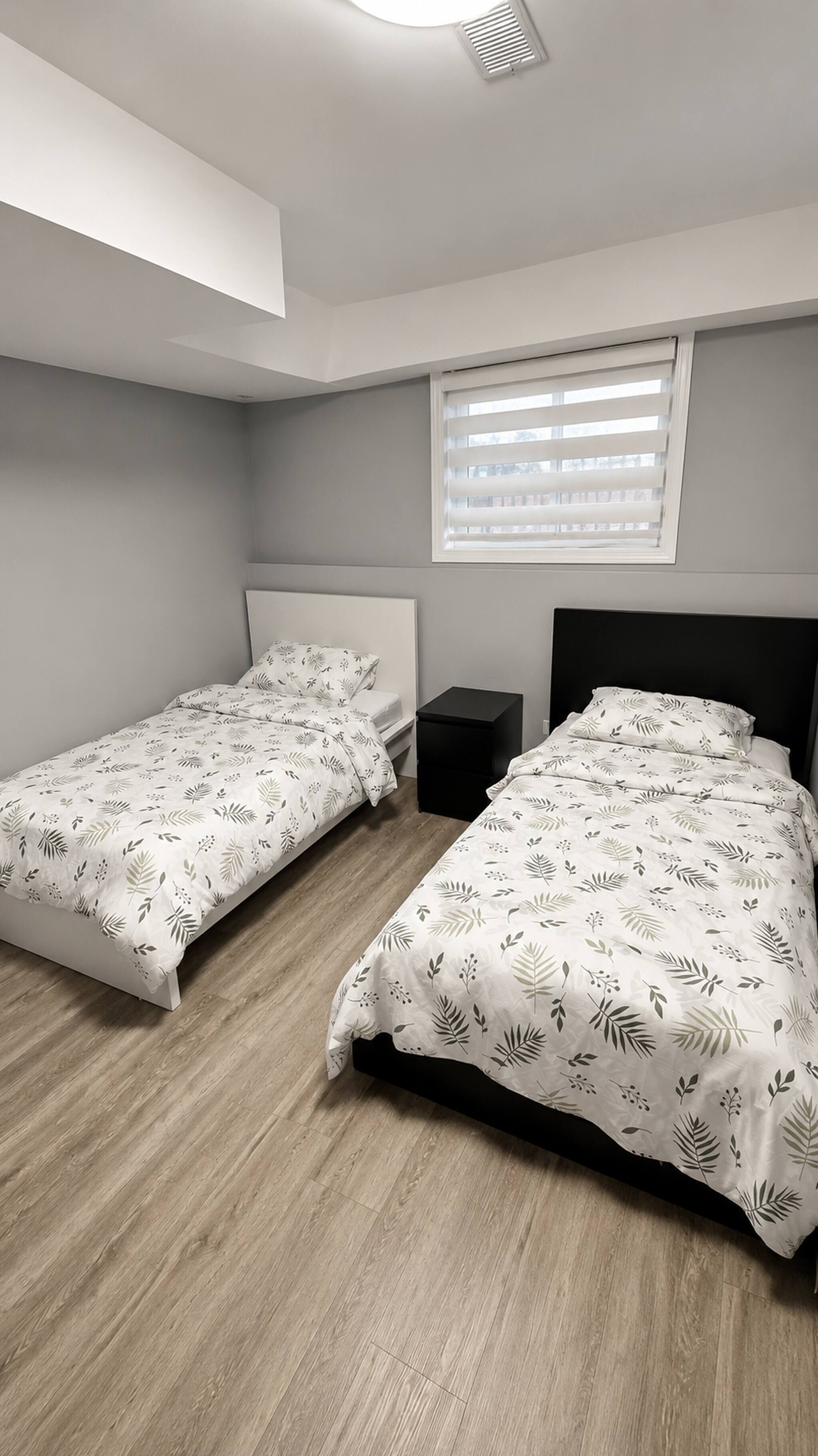 A bedroom with two beds, a window with blinds, a bedside table, and a ceiling vent.