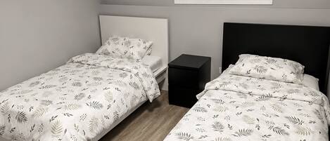 A bedroom with two beds, a window with blinds, a bedside table, and a ceiling vent.