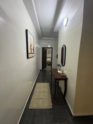 A hallway with a dark tiled floor, a beige rug, a wooden table with a mirror, and a framed picture on the wall. - STUNNING 2-Bedroom Apartment for Rent. (Dakar)