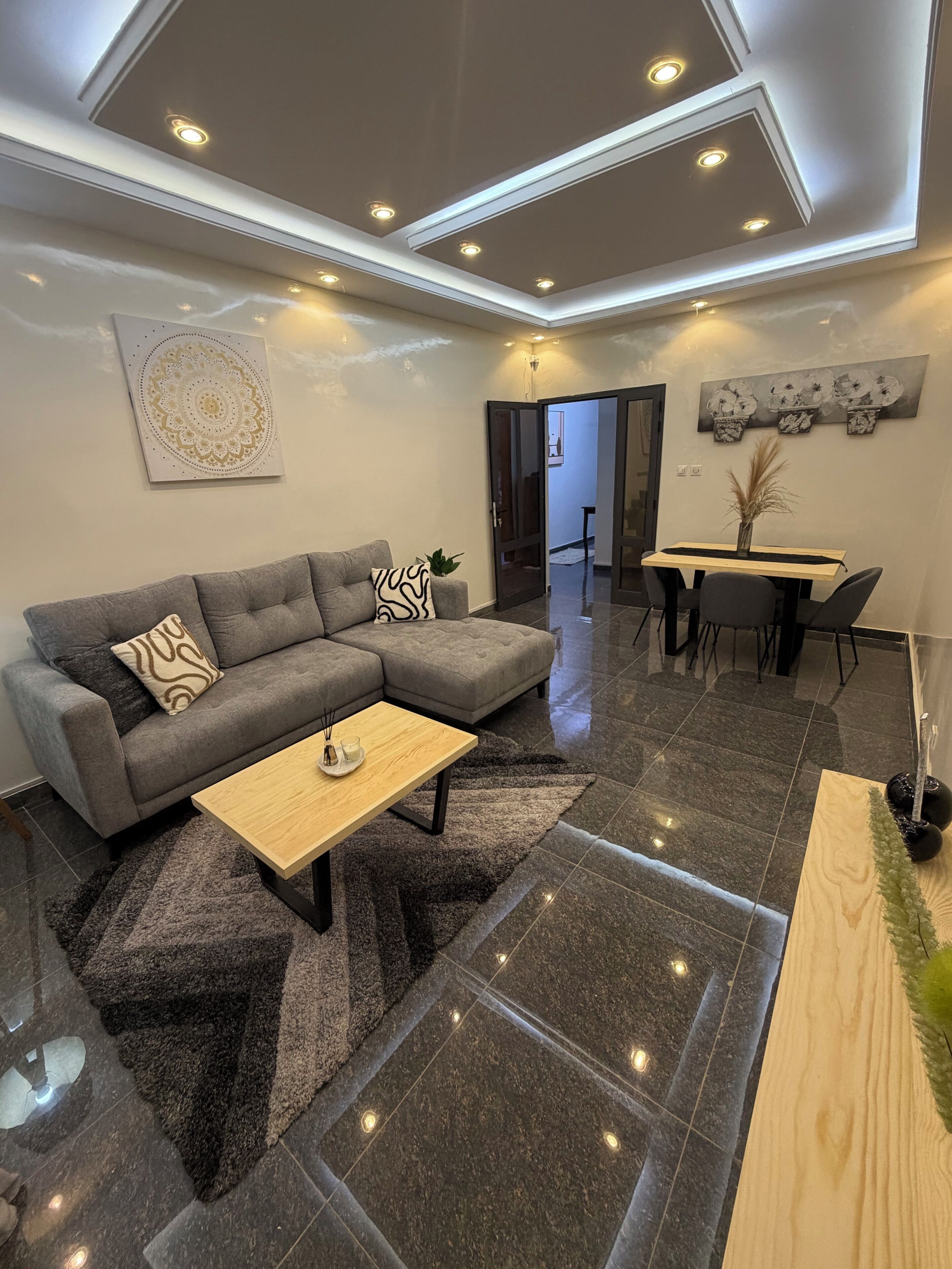 A modern living room with a grey sectional sofa, a wooden coffee table, a dining area with a wooden table and chairs, and a ceiling with recessed lighting.