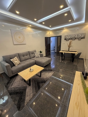 A modern living room with a grey sectional sofa, a wooden coffee table, a dining area with a wooden table and chairs, and a ceiling with recessed lighting. - STUNNING 2-Bedroom Apartment for Rent. (Dakar)