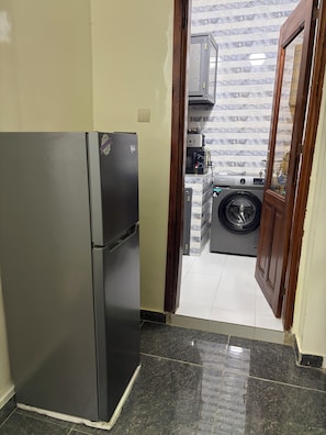 A kitchen with a refrigerator, washing machine, and a wooden door leading to a tiled area. - STUNNING 2-Bedroom Apartment for Rent. (Dakar)