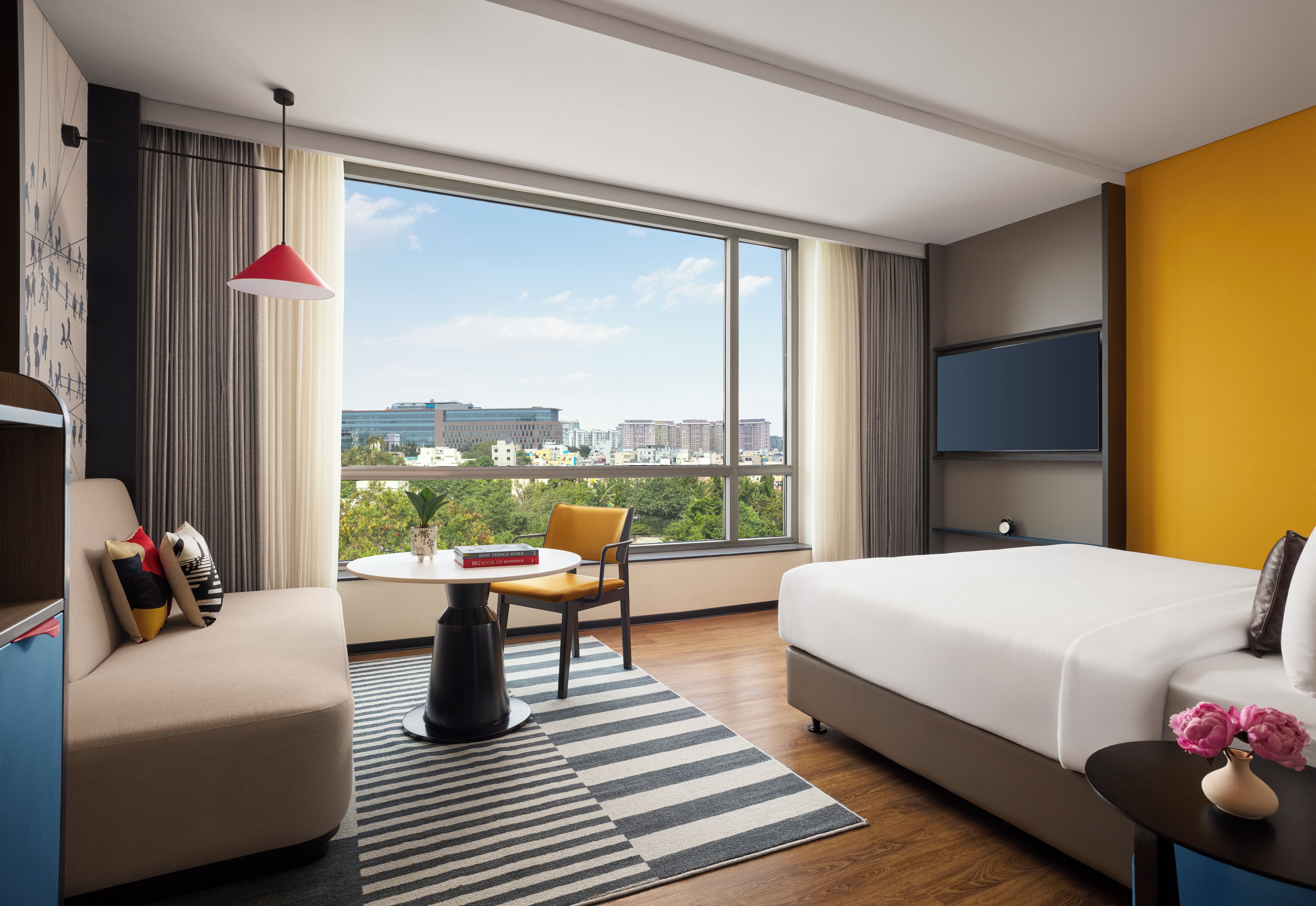 A hotel room with a bed, a desk, a chair, a TV, and a large window with a city view.
