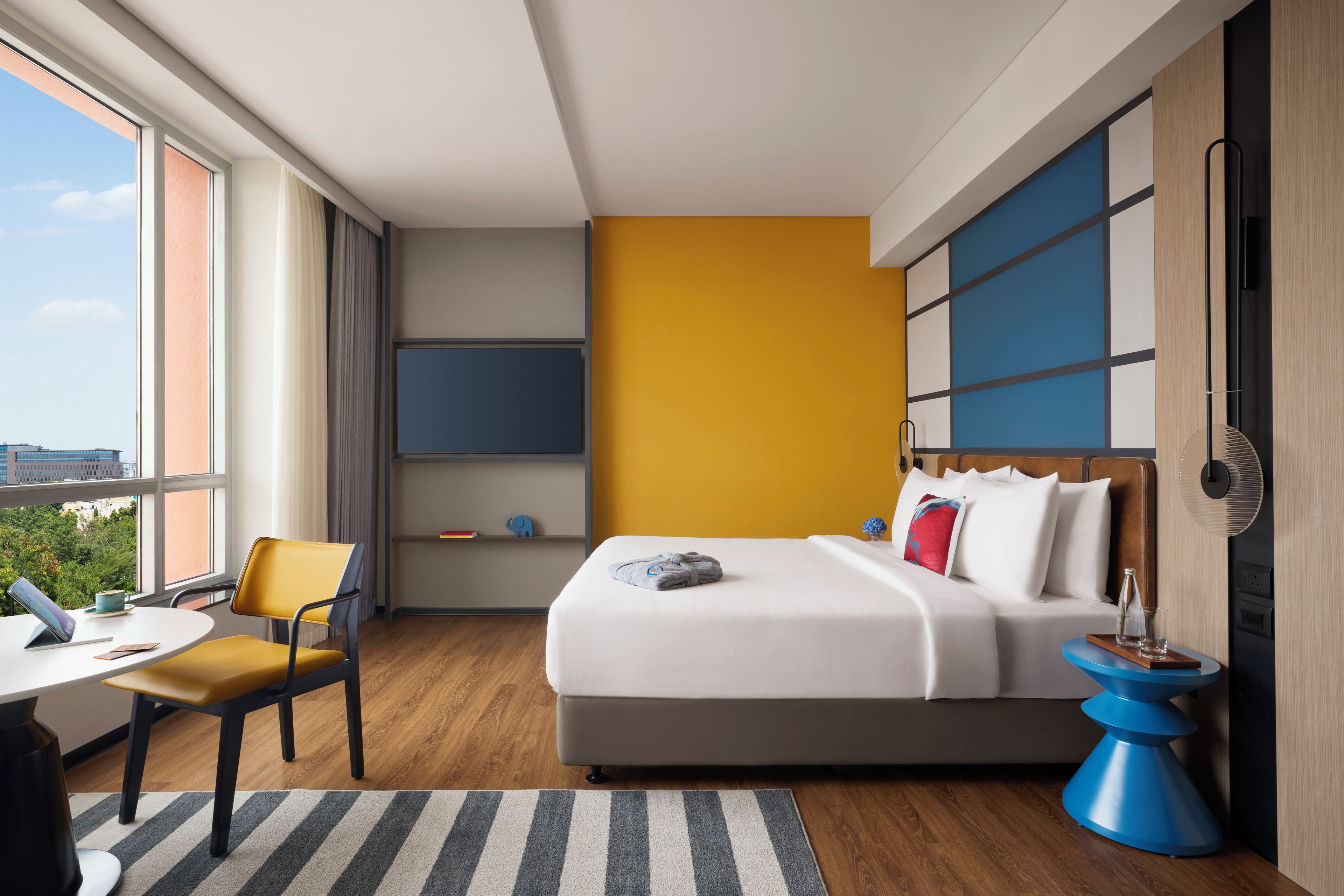 Habitación de hotel con cama, escritorio, silla, televisor y vista a la ciudad.