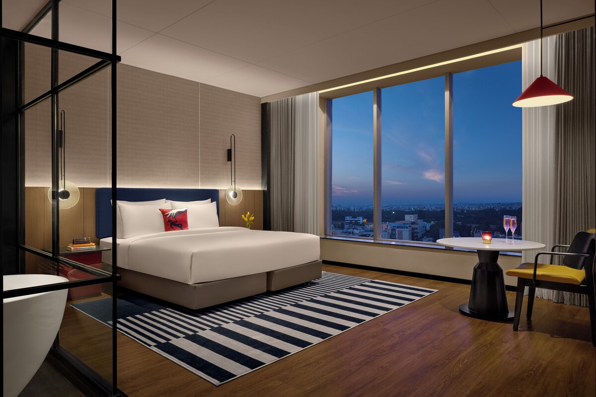 Habitación de hotel moderna con una cama grande, un área de descanso con una mesa y sillas, y vistas a la ciudad a través de amplios ventanales.