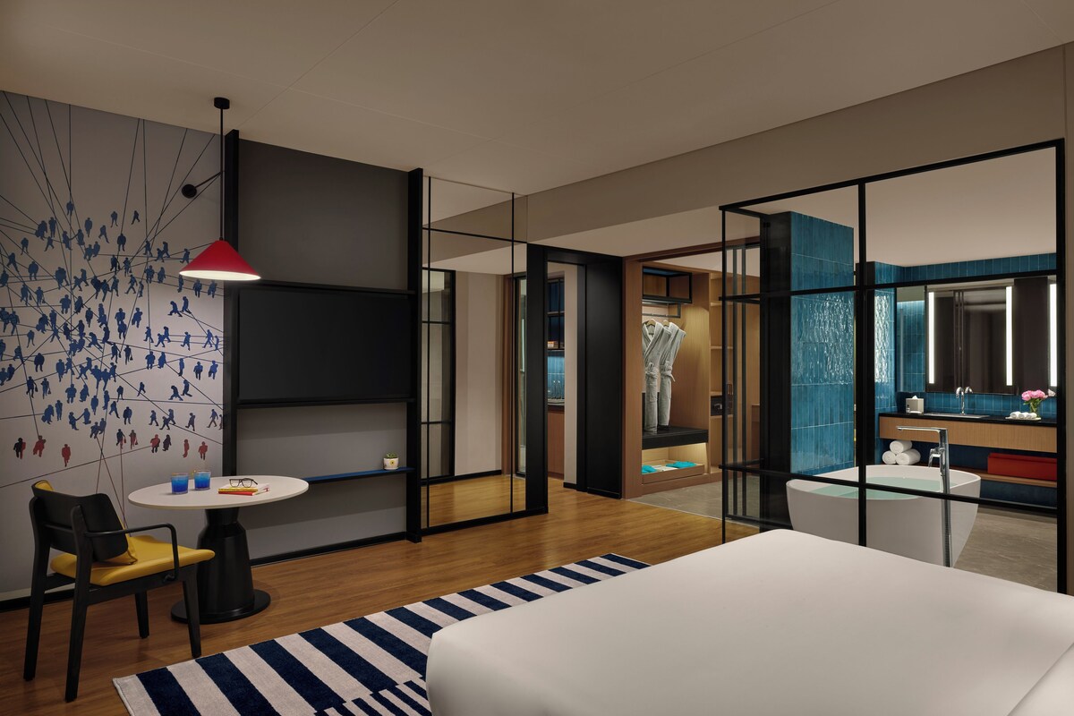 Habitación de hotel moderna con cama, escritorio, silla, mesa redonda, lámpara roja y una pared con alfombra azul y blanca a rayas.