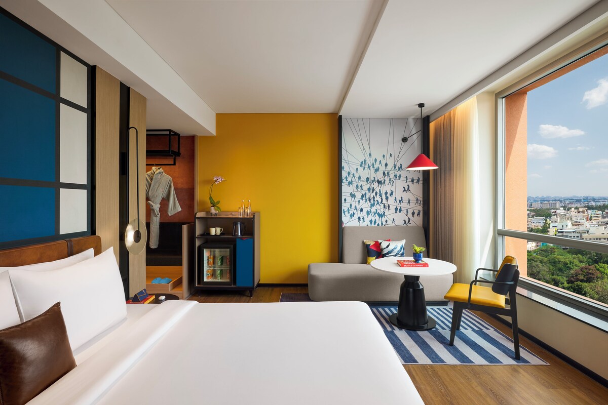 Una habitación de hotel con una cama grande, un pequeño espacio de descanso, un minibar y vistas a la ciudad.