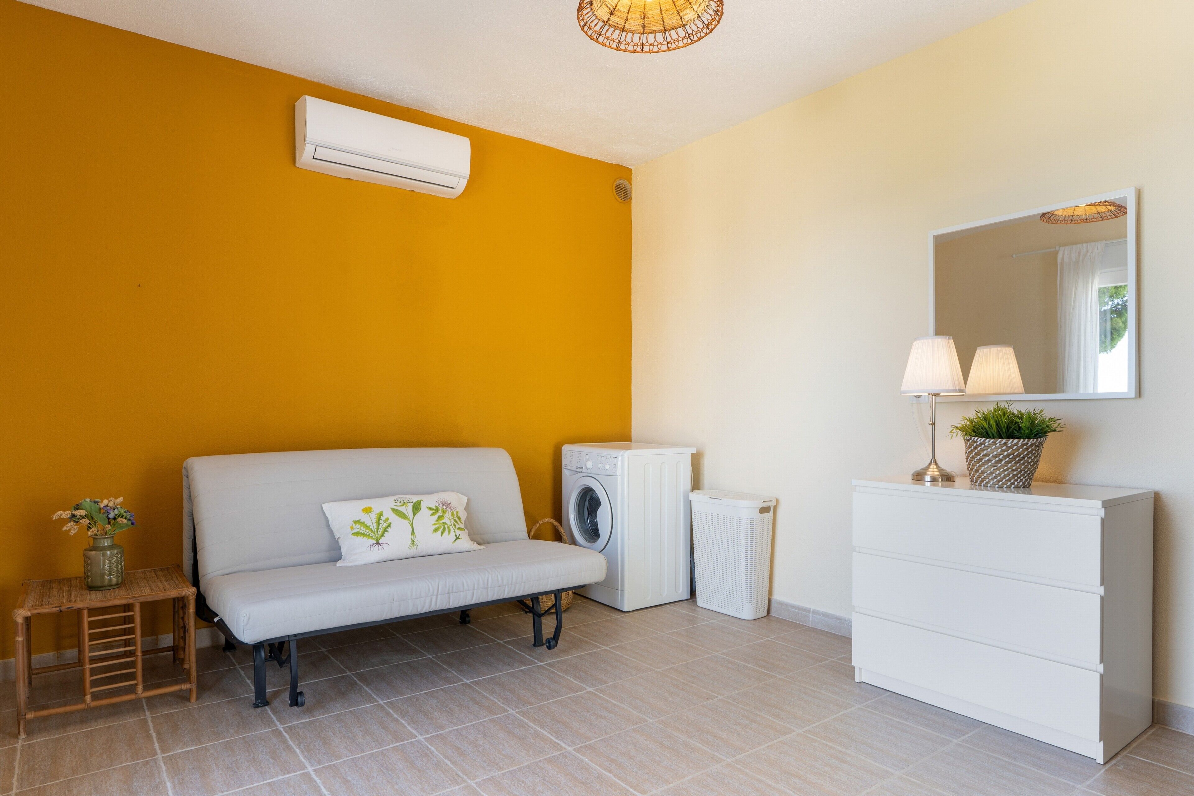 Una habitación con sofá, lavadora, secadora, una mesita con una planta y un aparador con espejo.