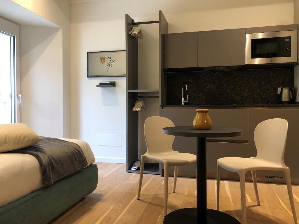 A modern room with a bed, a small round table with two chairs, a microwave, and a shelf with a framed picture.