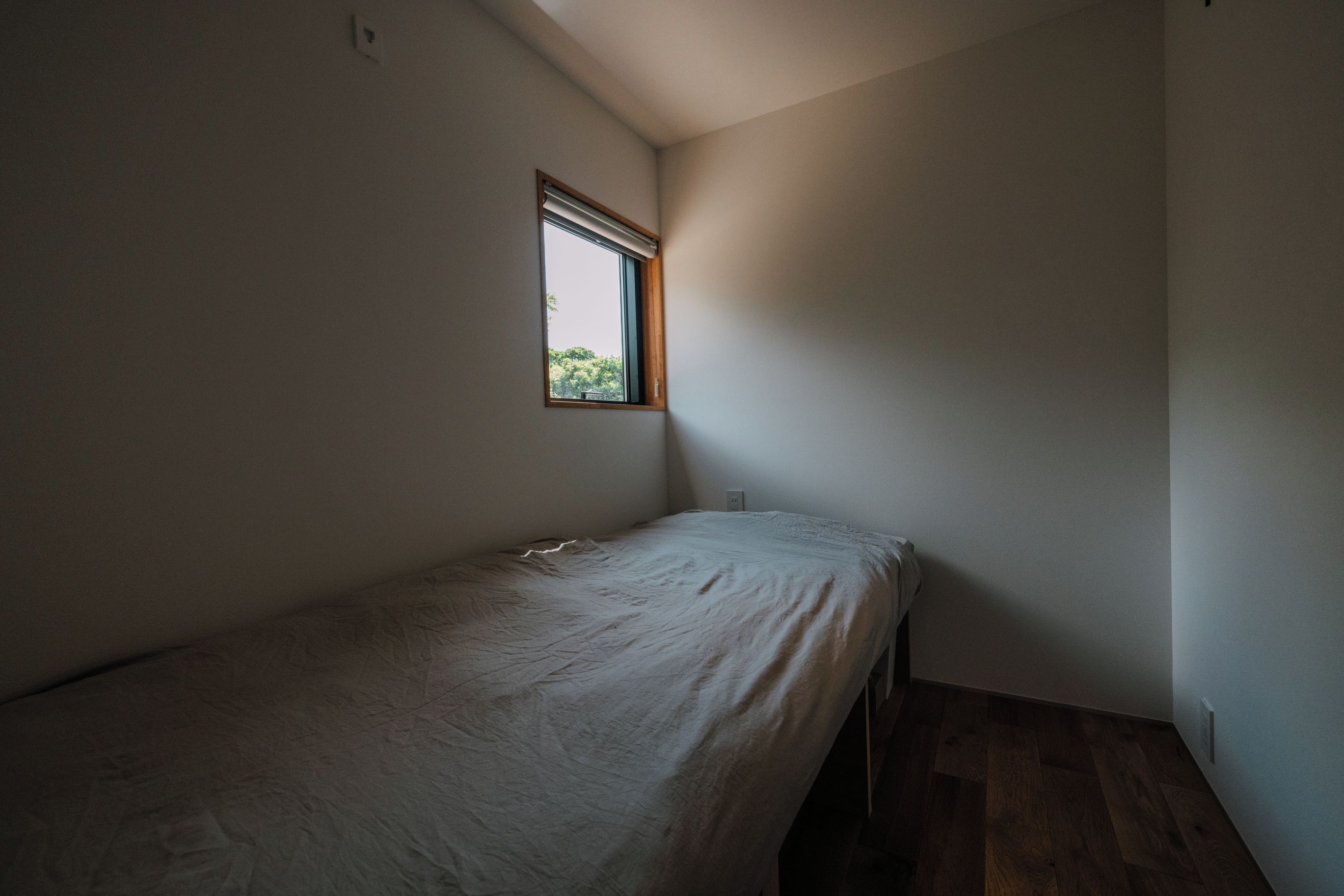 A small, dimly lit room with a single bed, a window, and a wooden floor.
