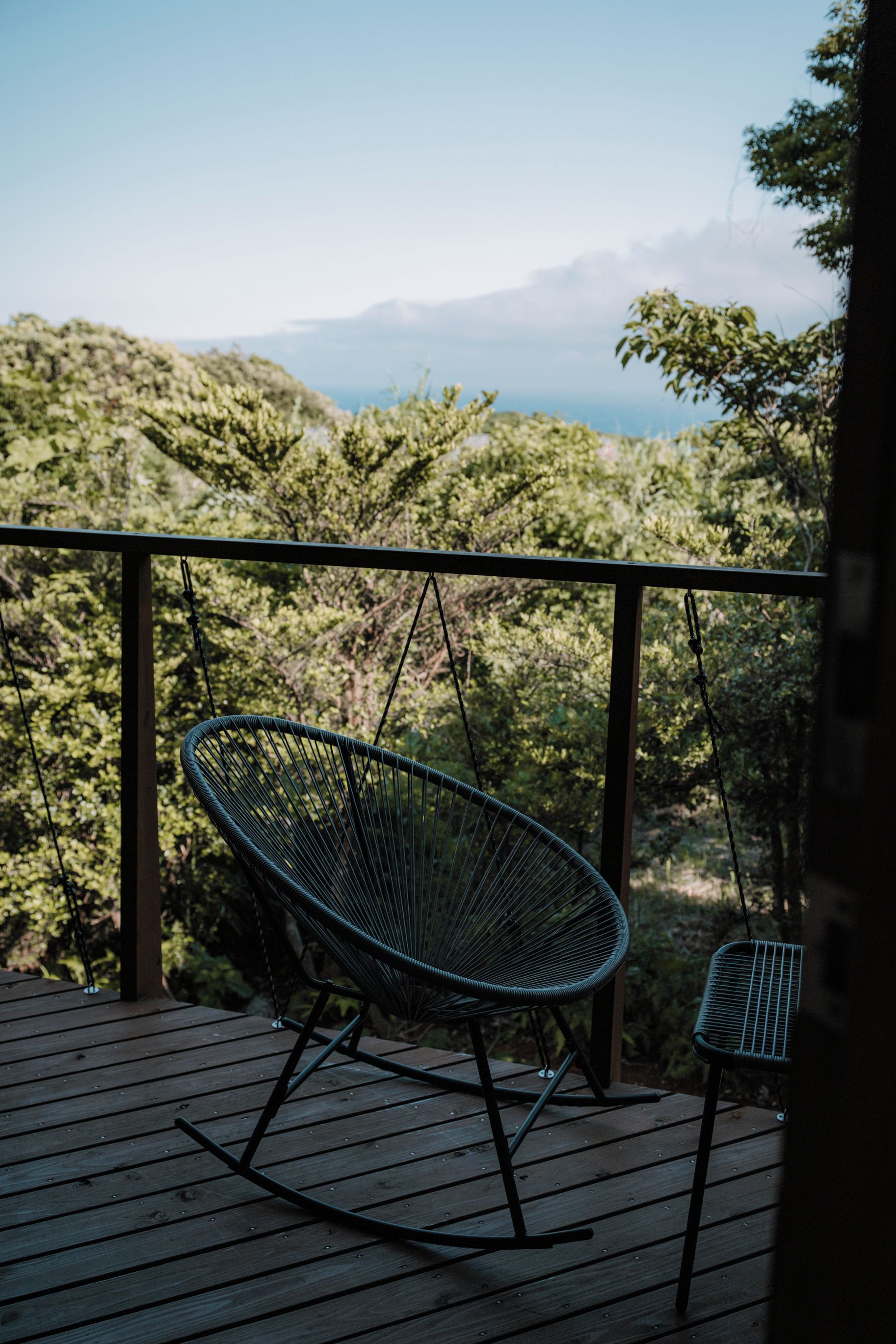 Eine Holzterrasse mit einem Metallschaukelstuhl und einem kleineren Stuhl, mit Blick auf eine gebirgige Landschaft.