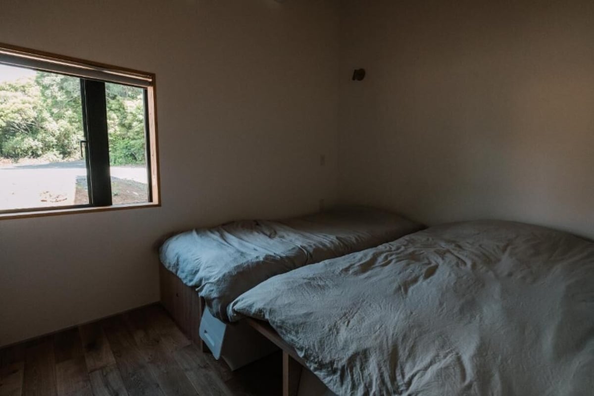 Una habitación pequeña con cama individual, ventana, suelo de madera y una cama sencilla.