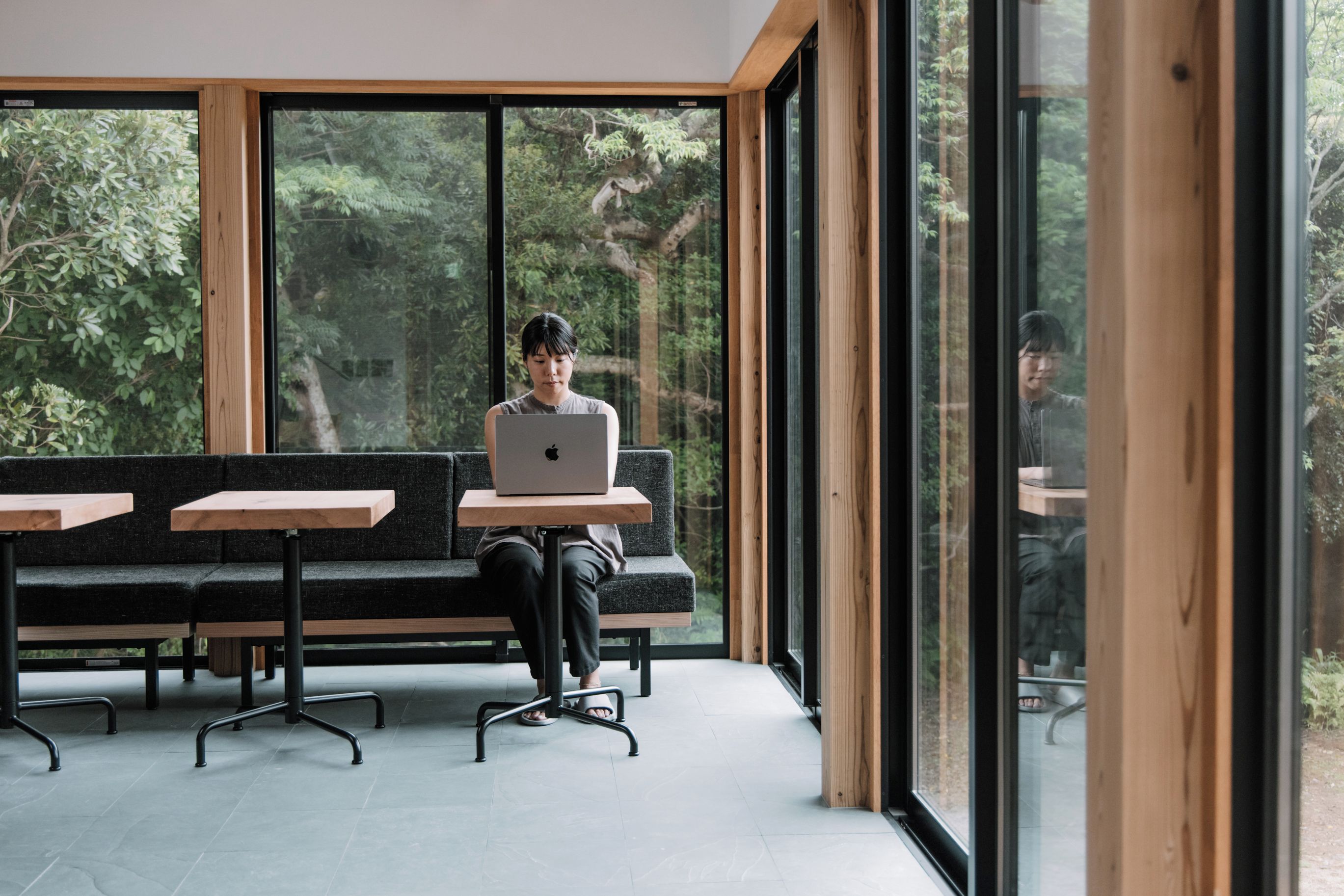 A person seated on a bench with a laptop, in a room with large windows and a view of trees.
