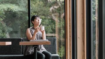 A person sitting on a bench with a laptop and a cup, in a room with large windows and a view of trees.