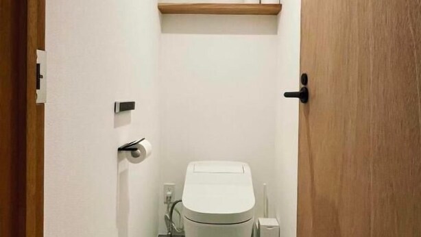 Un baño estrecho con inodoro, un estante de madera arriba y una puerta a la derecha.