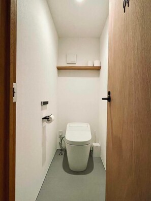 A narrow bathroom with a toilet, a wooden shelf above, and a door on the right. - Arc yakushima (Yakushima)