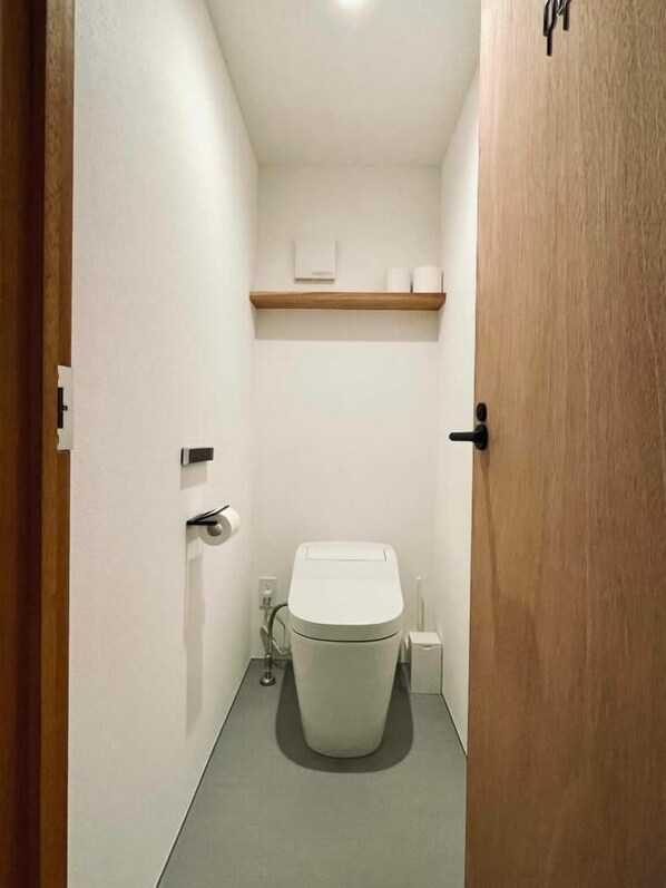 Un baño estrecho con inodoro, un estante de madera arriba y una puerta a la derecha.