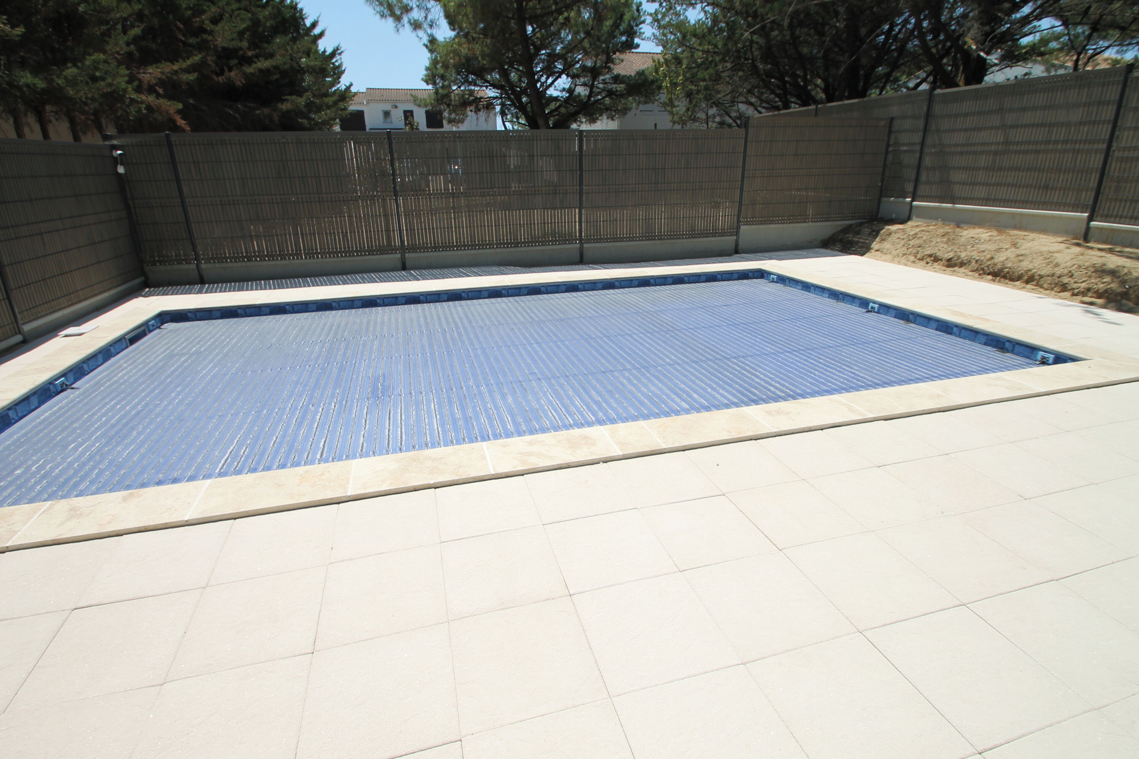 A rectangular swimming pool with a blue interior, surrounded by a tiled deck and a dark fence.