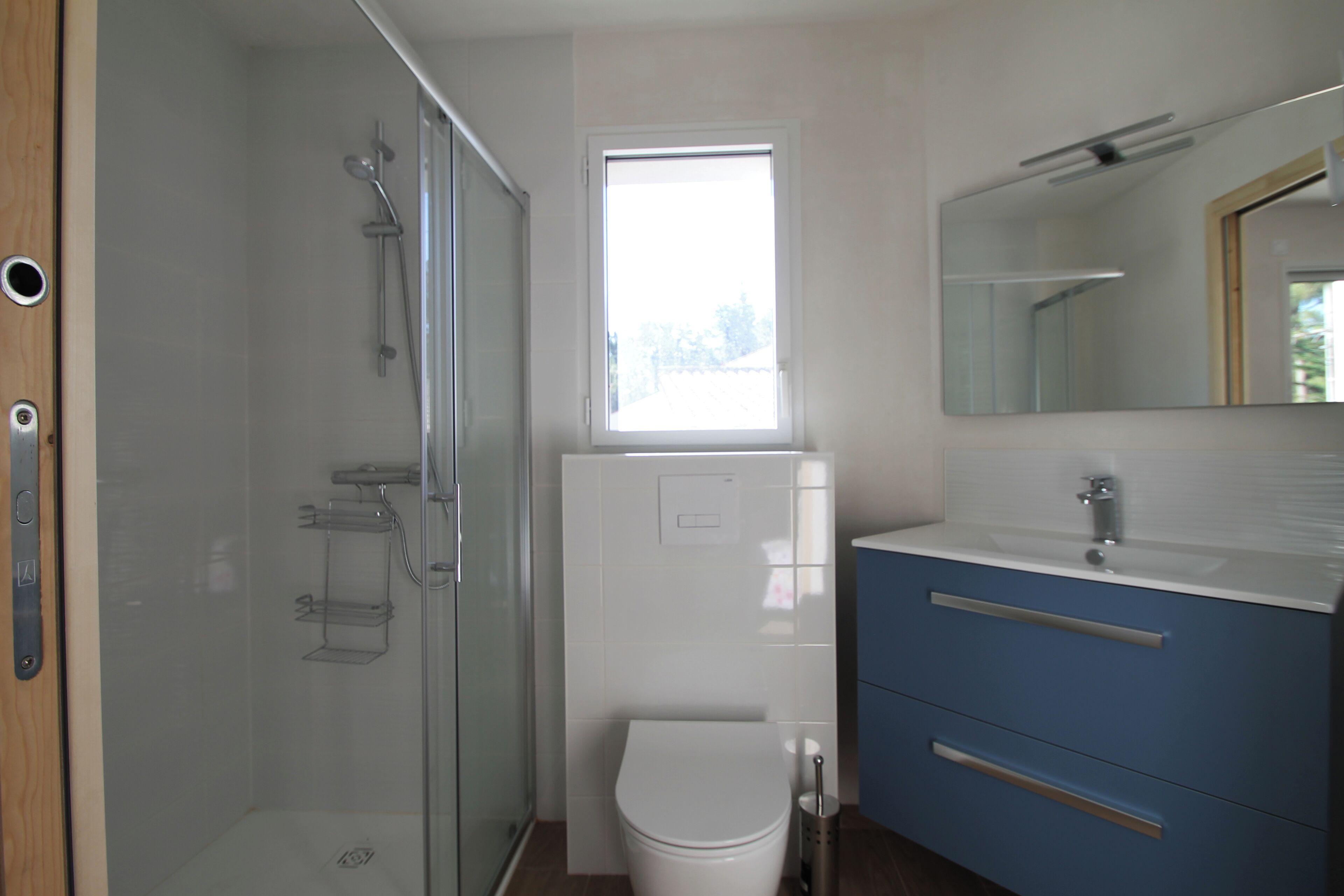 A bathroom with a glass shower enclosure, a white toilet, a sink with a mirror, and a window providing natural light.
