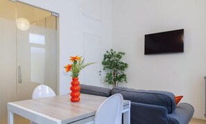 Ein modernes Wohnzimmer mit einer Couch, einem Esstisch mit Stühlen, einem an der Wand befestigten Fernseher und einer Zimmerpflanze.