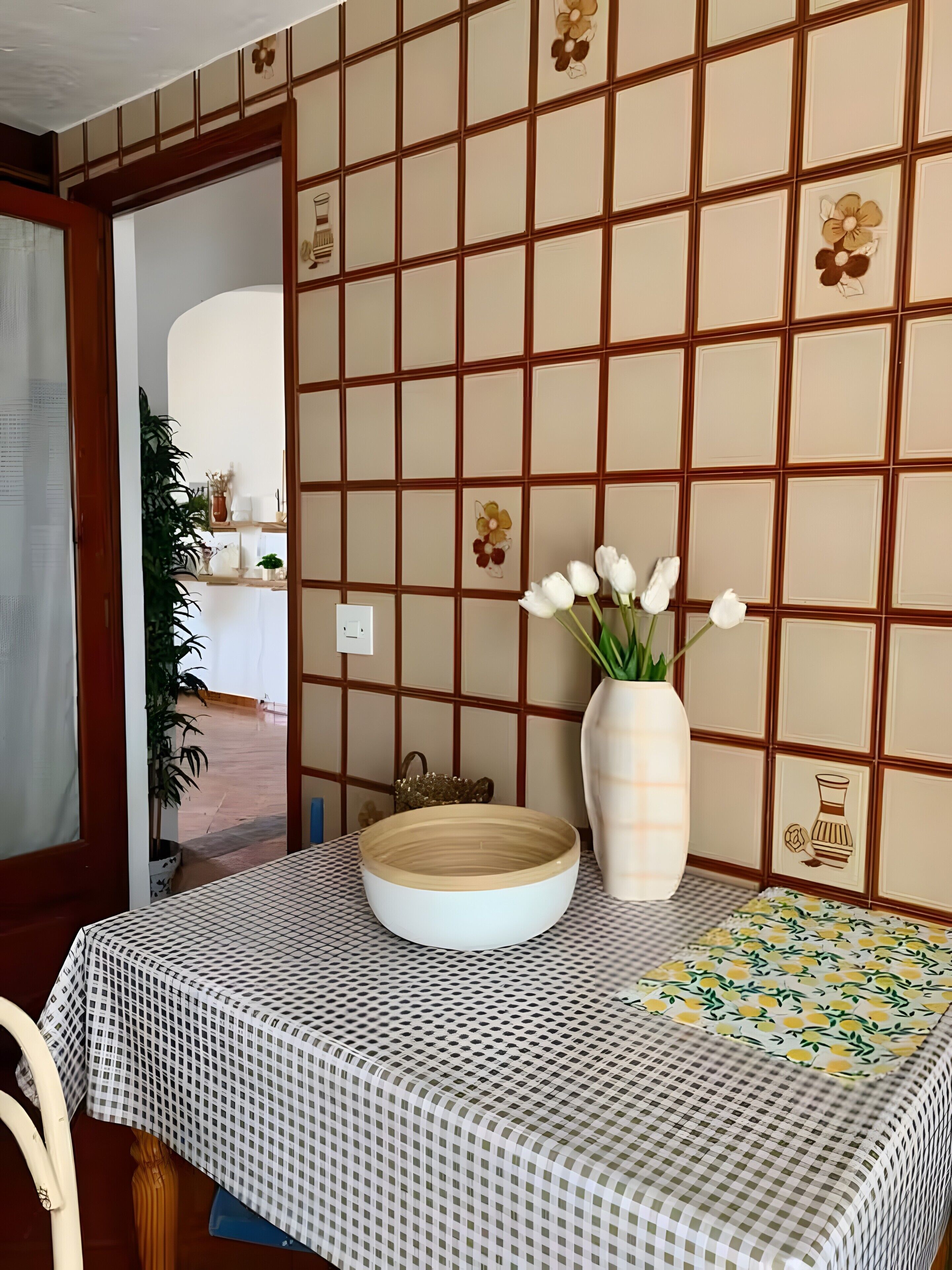 A dining area with a checkered tablecloth, a vase with white flowers, and a decorative wall with a grid pattern and floral motifs.