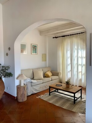 A living room with a sofa, a coffee table, a lamp, a plant, and a window with curtains.