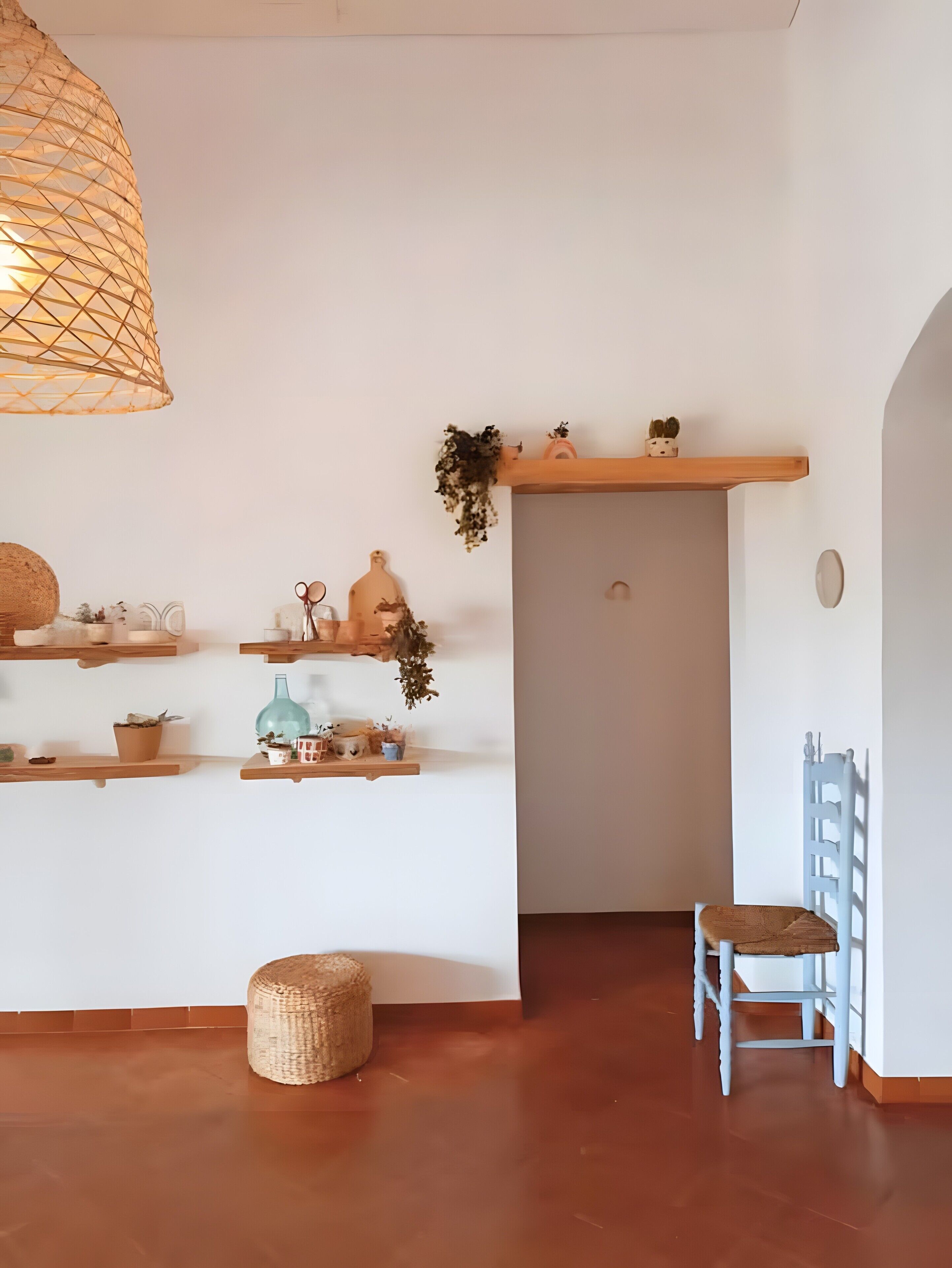 A room with a woven light fixture, wooden shelves with various items, a small round ottoman, and a chair.