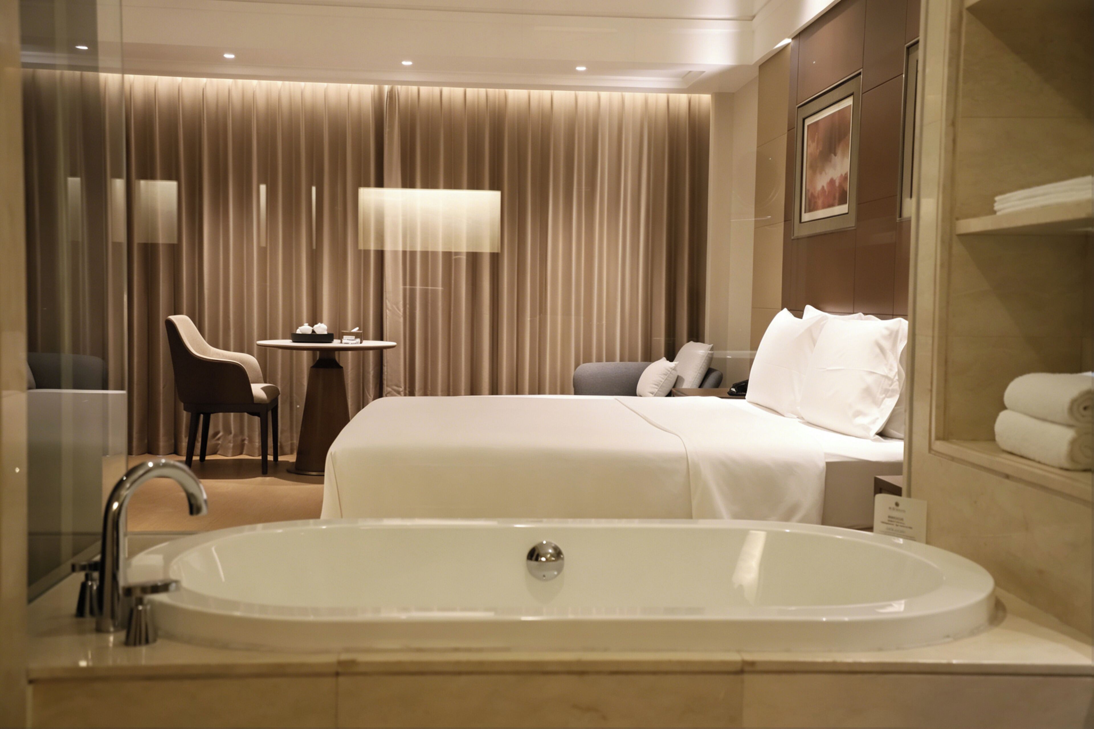 Habitación de hotel con una bañera grande, una cama con almohadas, una silla, una mesita con una lámpara y un espejo.