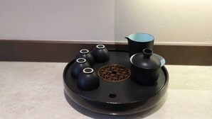 Una mesa negra y redonda de diseño moderno con un motivo decorativo central, rodeada por cuatro jarrones negros y una jarrilla negra.