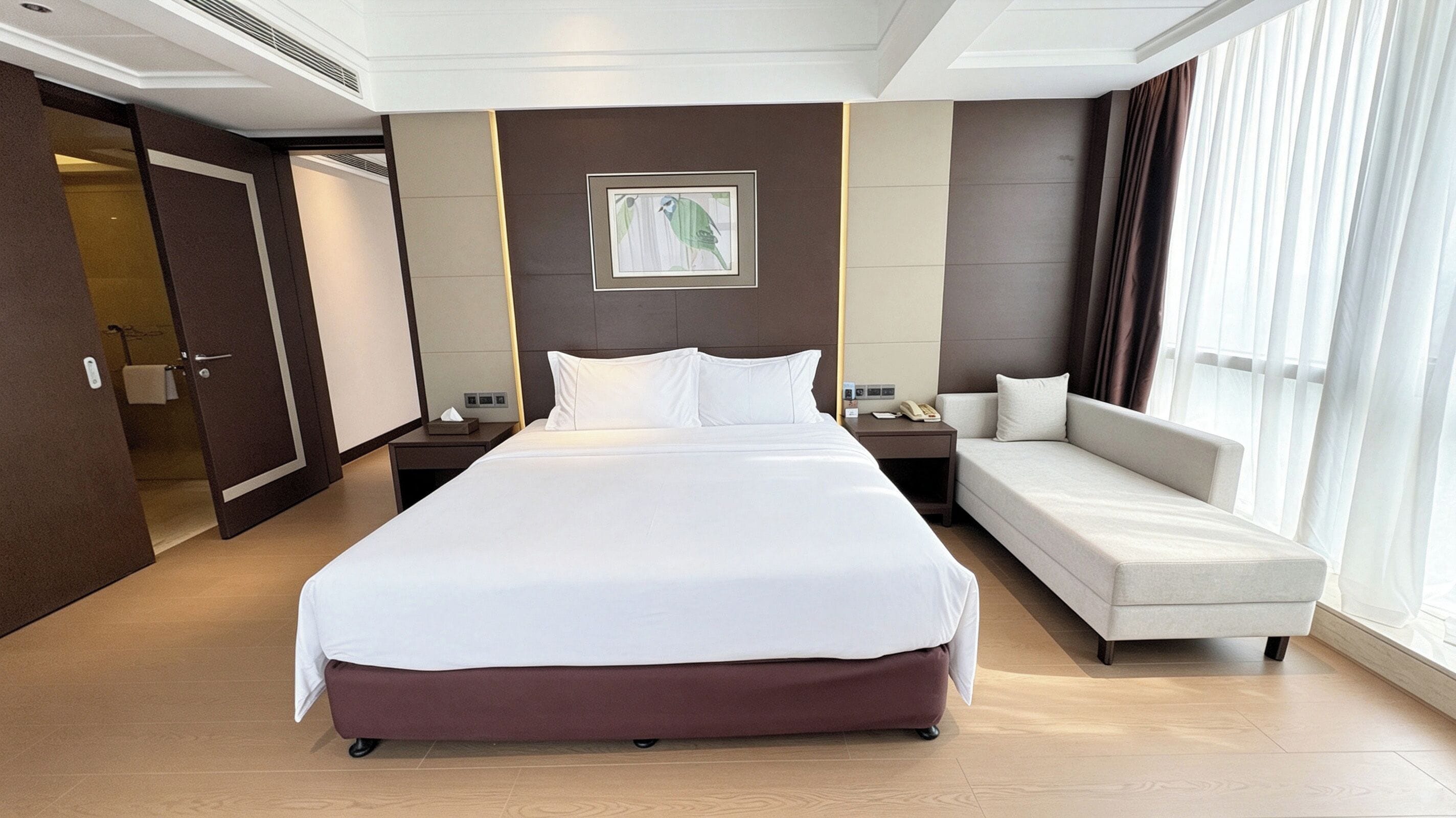A hotel room with a large bed, a sofa, a bedside table, and a window with curtains.