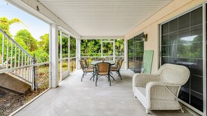 Een overdekt terras met een tafel, stoelen en een rieten stoel, met uitzicht op groen.