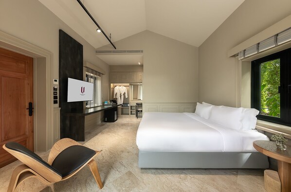 A hotel room with a large bed, a wooden chair, a desk with a computer, and a window with a view of greenery.