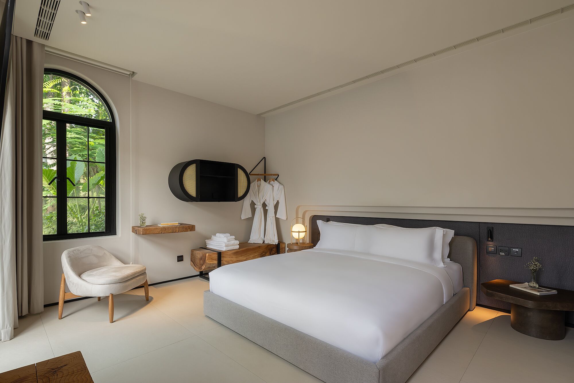 Una habitación de hotel moderna con una cama grande, una zona de estar con una silla, una mesita de noche con una lámpara y vistas a zonas verdes a través de una ventana.
