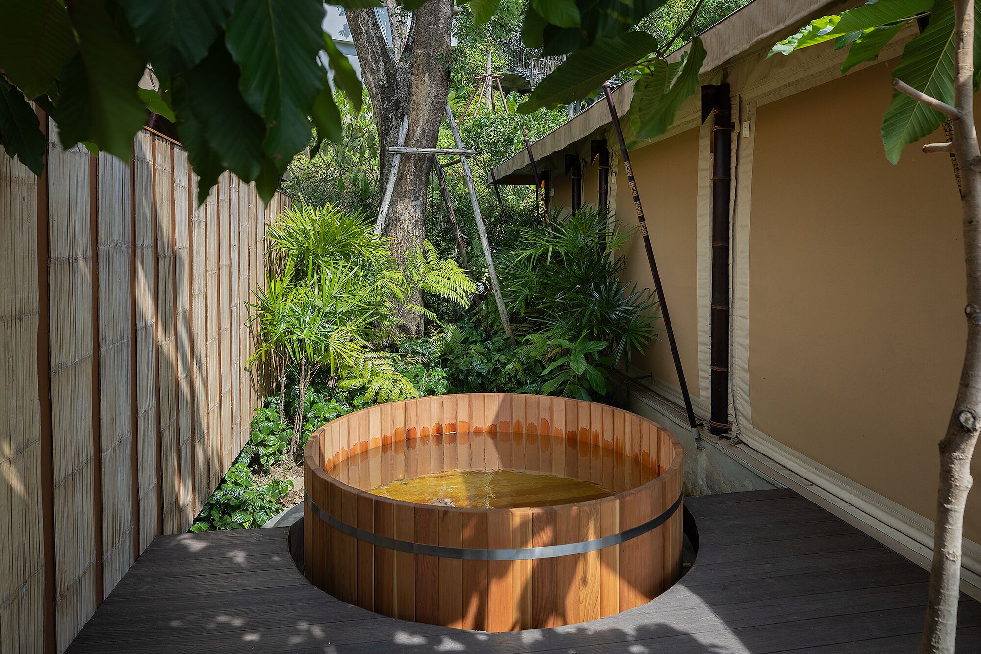 A wooden hot tub on a deck with a bamboo fence and tropical plants in the background.