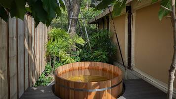 A wooden hot tub on a deck with a bamboo fence and tropical plants in the background.