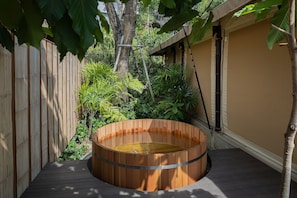 Un bain à remous en bois installé sur une terrasse, avec une clôture en bambou et des plantes tropicales en arrière-plan.