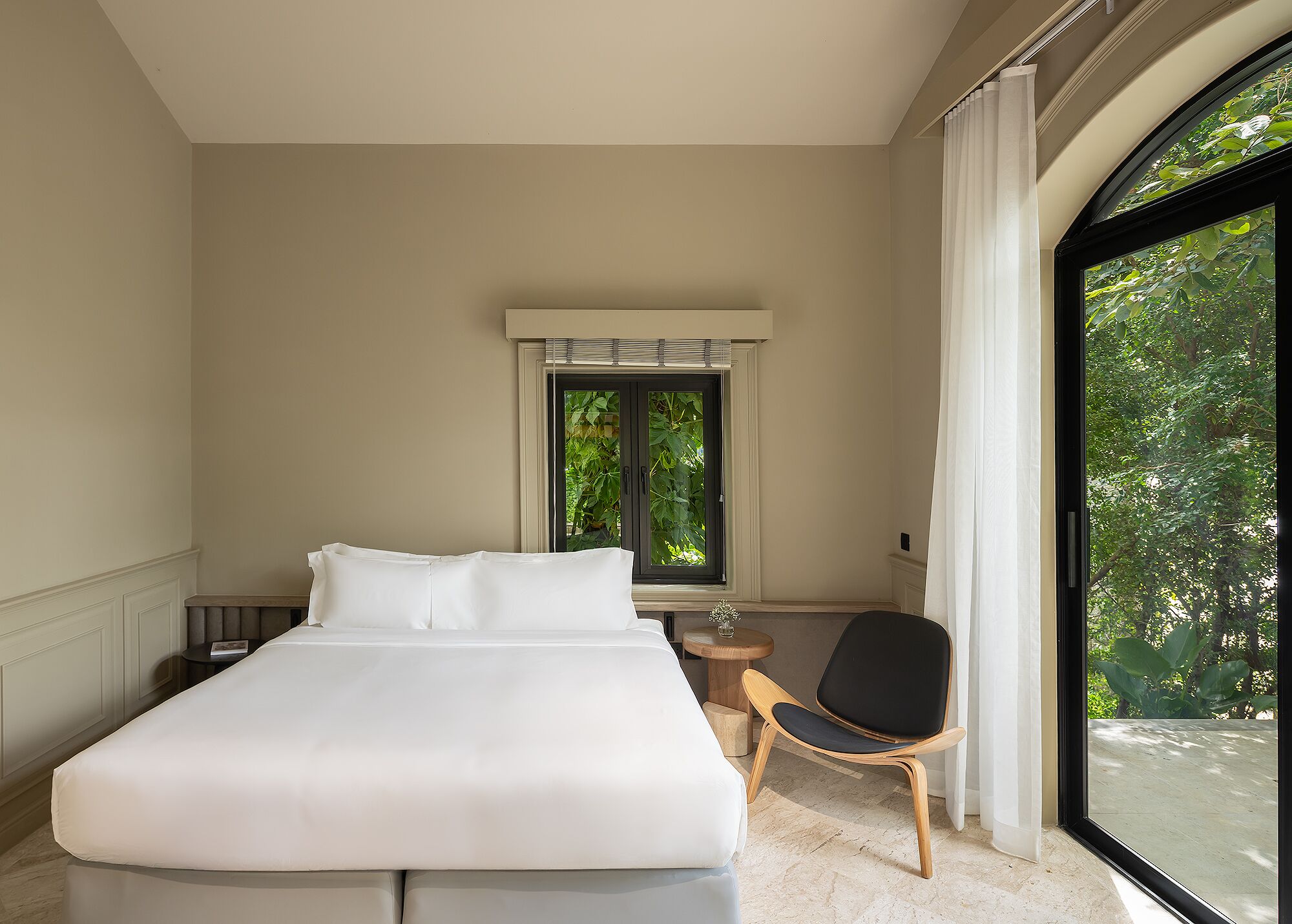 Un dormitorio con una cama grande, una silla, una mesita y un ventanal con vistas a zonas verdes.