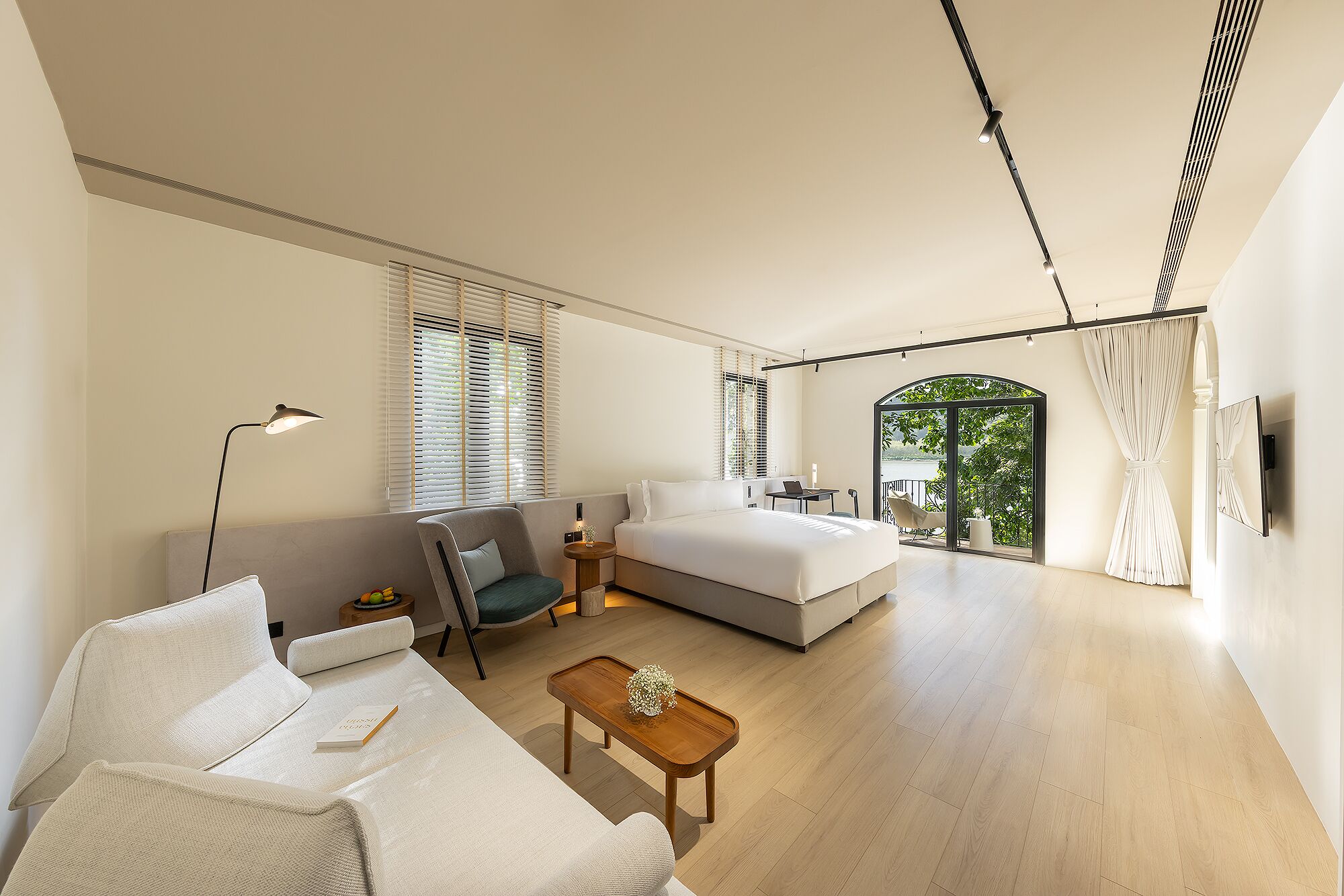 A modern bedroom with a large bed, a sofa, a small table, and a desk. There is a window with blinds, a door leading to a balcony, and a ceiling with track lighting.