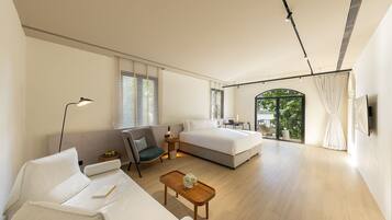 A modern bedroom with a large bed, a sofa, a small table, and a desk. There is a window with blinds, a door leading to a balcony, and a ceiling with track lighting.