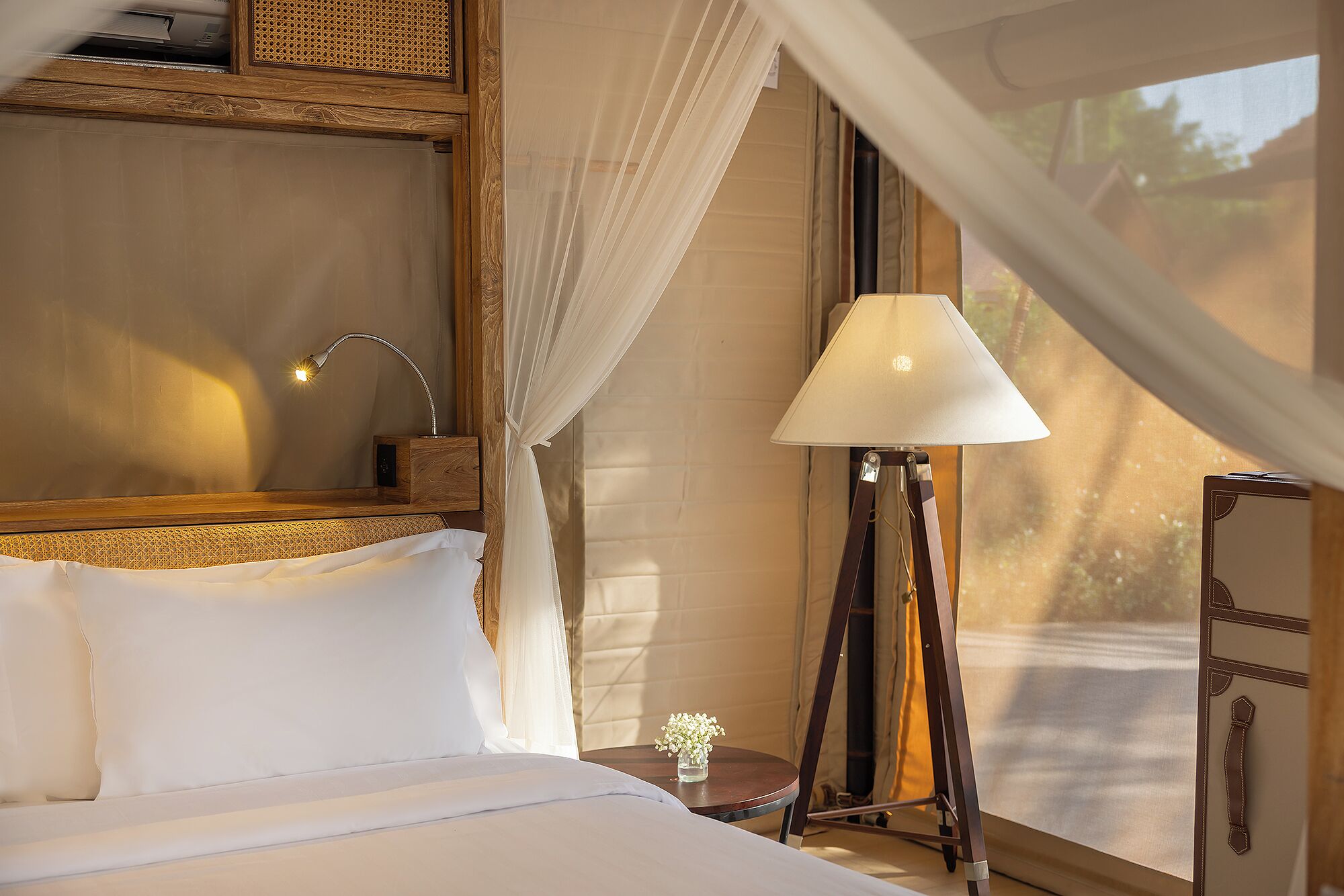 A neatly made bed with white linens, a bedside lamp, a small table with a vase of flowers, and a window with sheer curtains.