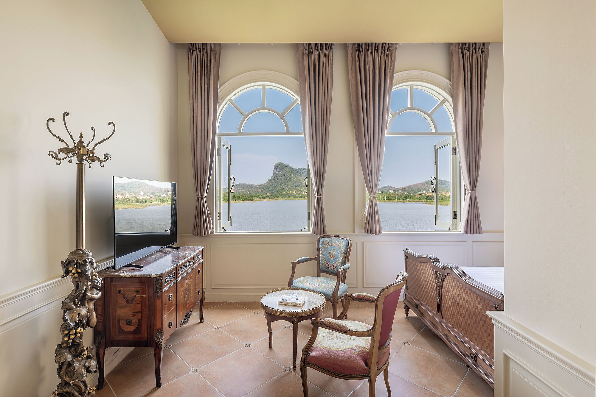 A room with a TV, a wooden cabinet, two chairs, a round table, and large windows with curtains.